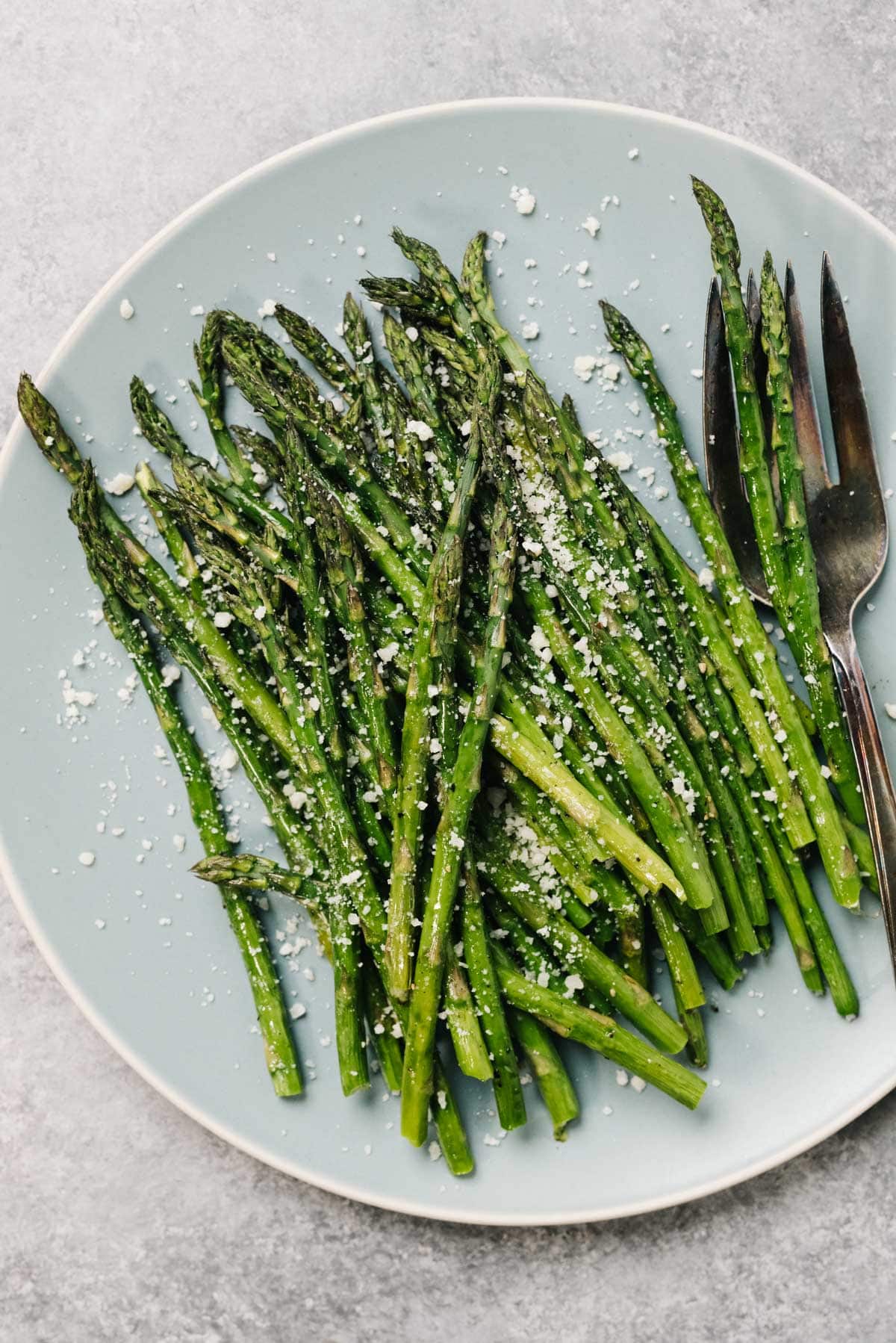 Roasted asparagus spears on a blue plate, garnished with grated parmesan cheese, with a silver serving fork.