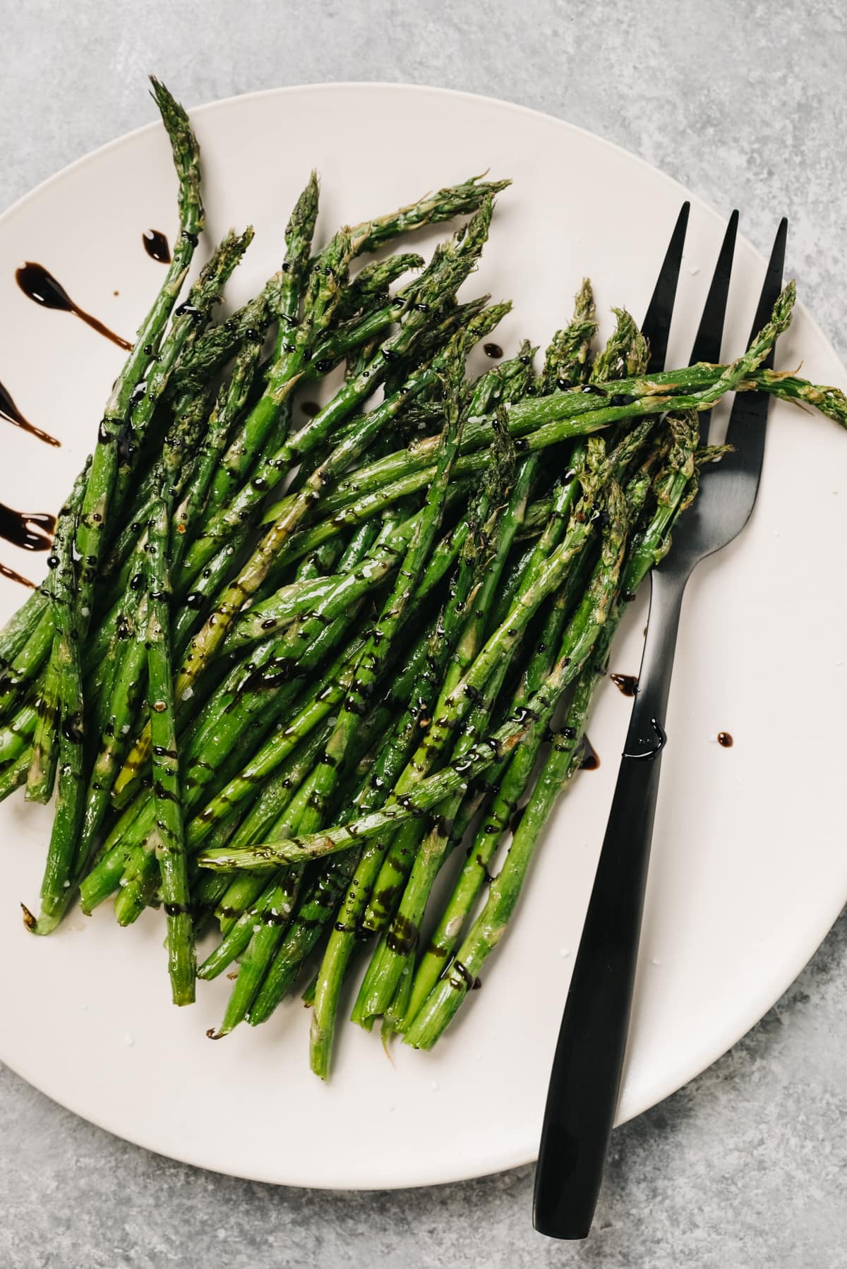 Cooked asparagus spears on a white platter, drizzled with balsamic reduction, with a black serving fork.