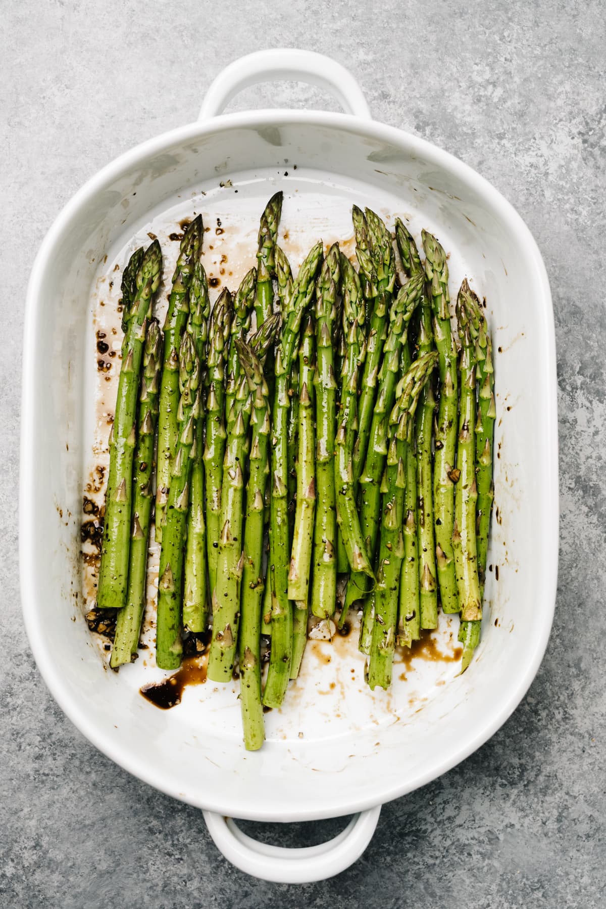 Asparagus spears in a casserole dish with olive oil, balsamic vinegar, salt, and pepper.