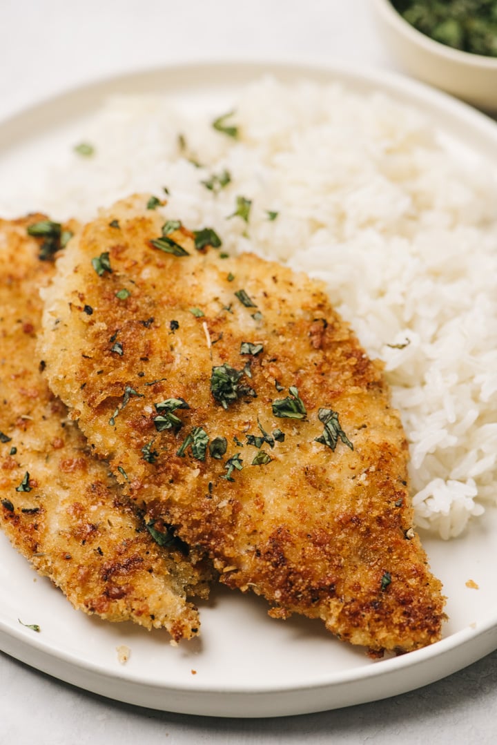 Side view, two golden brown parmesan crusted chicken breasts over white rice on a white place, with a small bowl of fresh chopped parsley in the background.