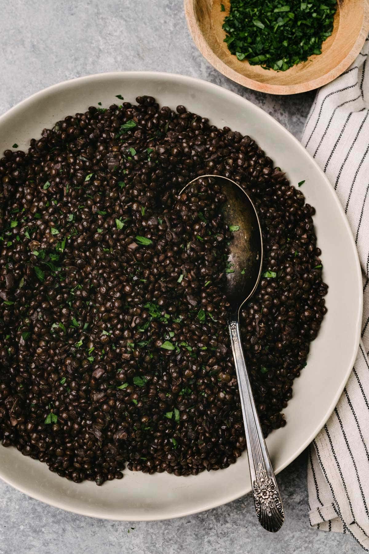 A silver serving spoon tucked into a tan serving bowl filled with black lentils; a striped linen napkin and small wood bowl of chopped parsley are to the side of the bowl.