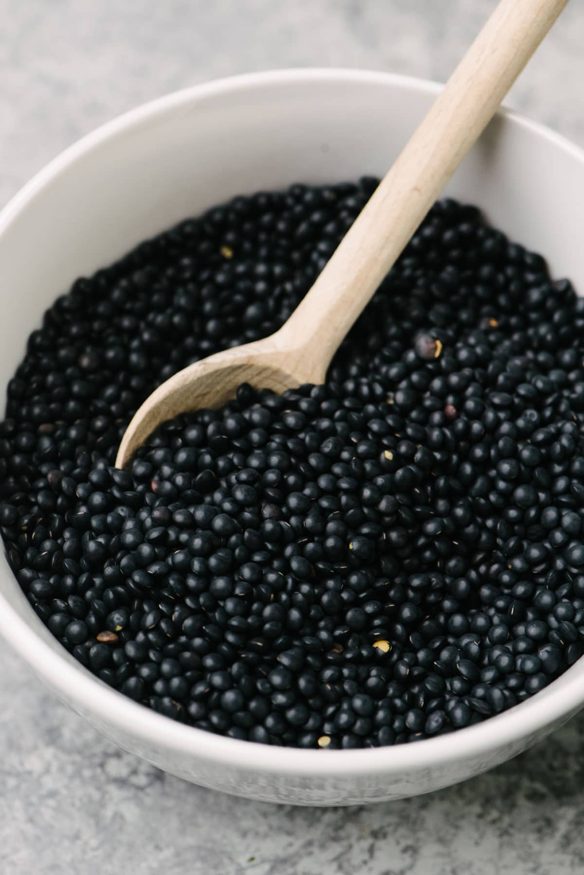 Side view, a small wood spoon tucked into a white bowl of dried black beluga lentils.