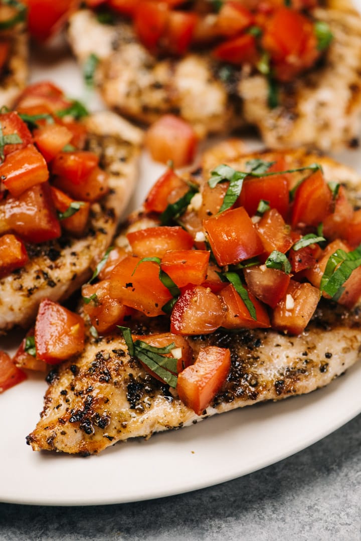 Side view, grilled chicken breasts topped with tomato bruschetta on a cream serving platter.