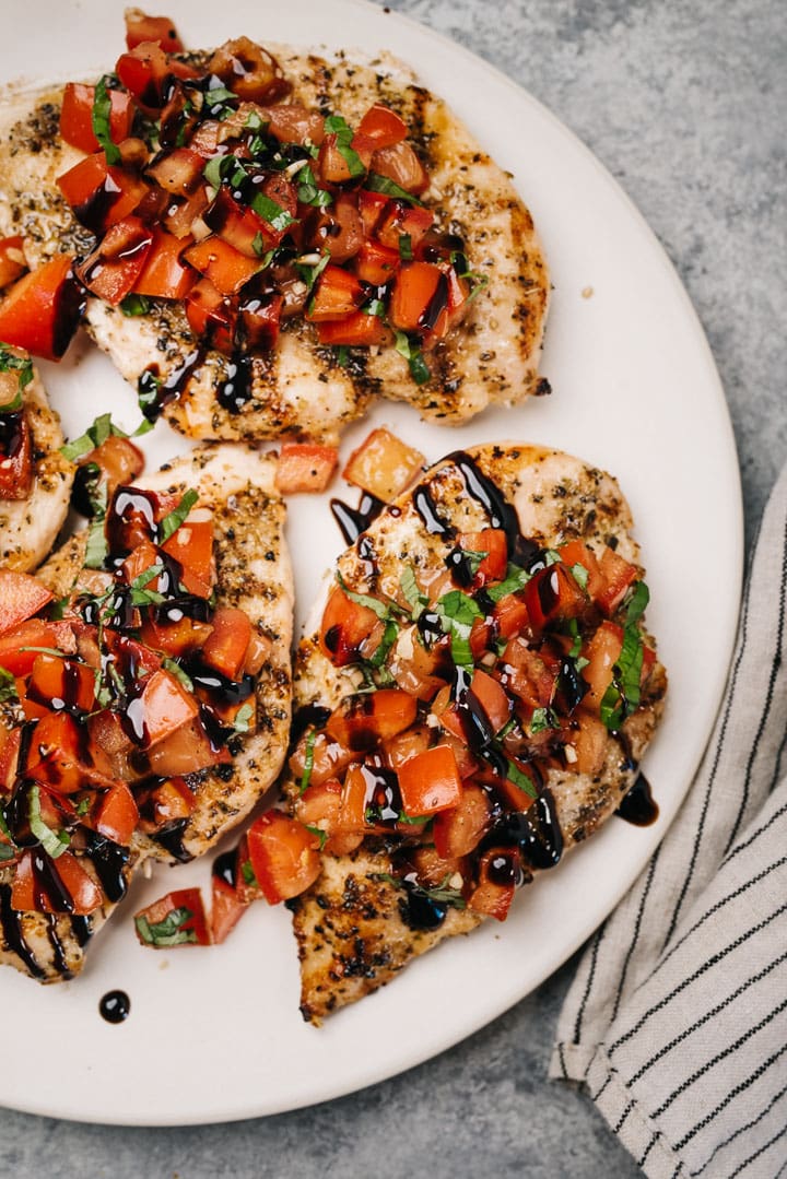 Three grilled chicken breasts topped with tomato bruschetta and garnished with balsamic glaze on a cream serving platter with a striped linen napkin.