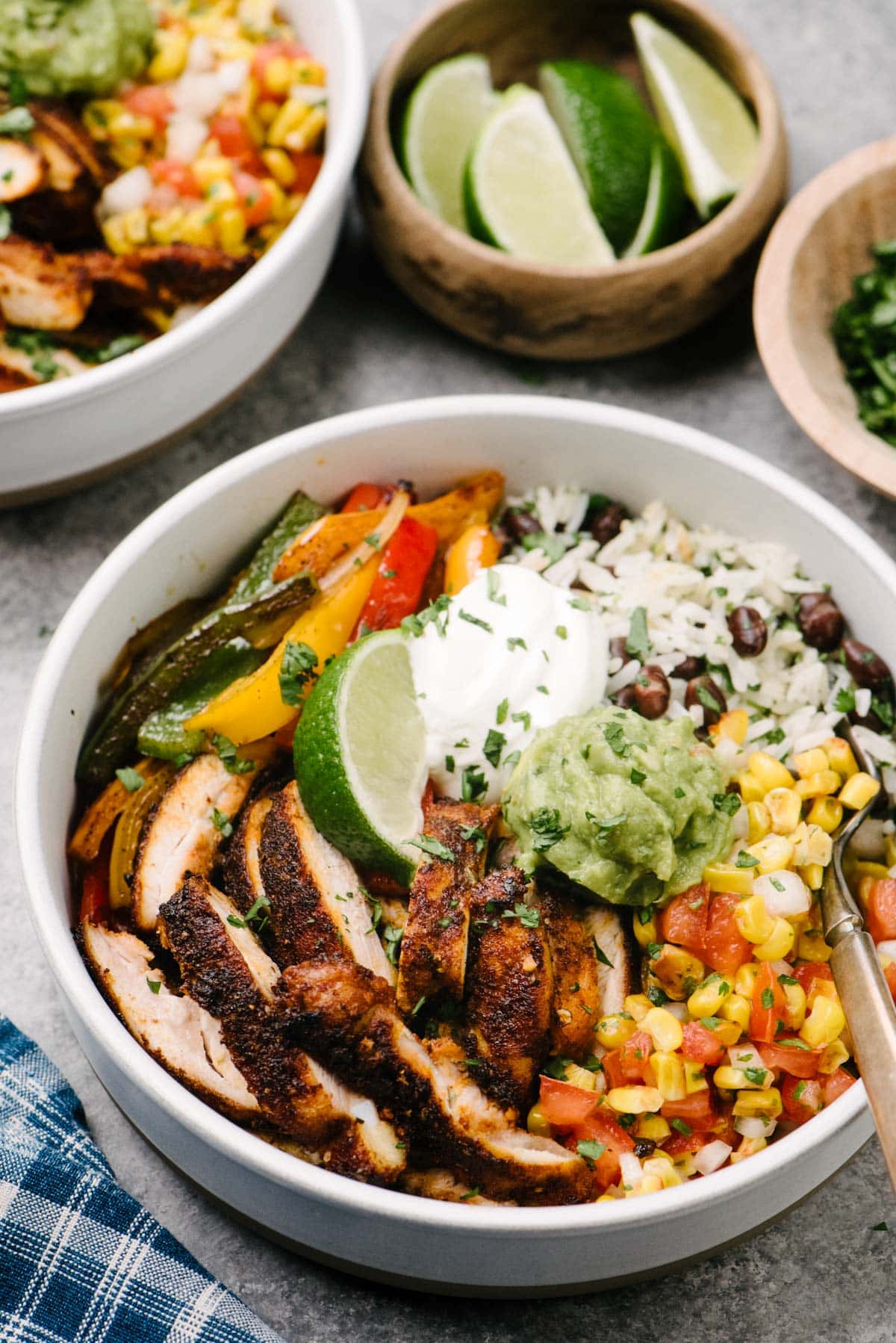 Side view - a chicken fajita bowl with seared chicken, onions and bell peppers, black beans and rice, and corn salsa, topped with guacamole, sour cream, and a lime wedge. A second fajita bowl and small bowls of lime wedges and cilantro are in the background.