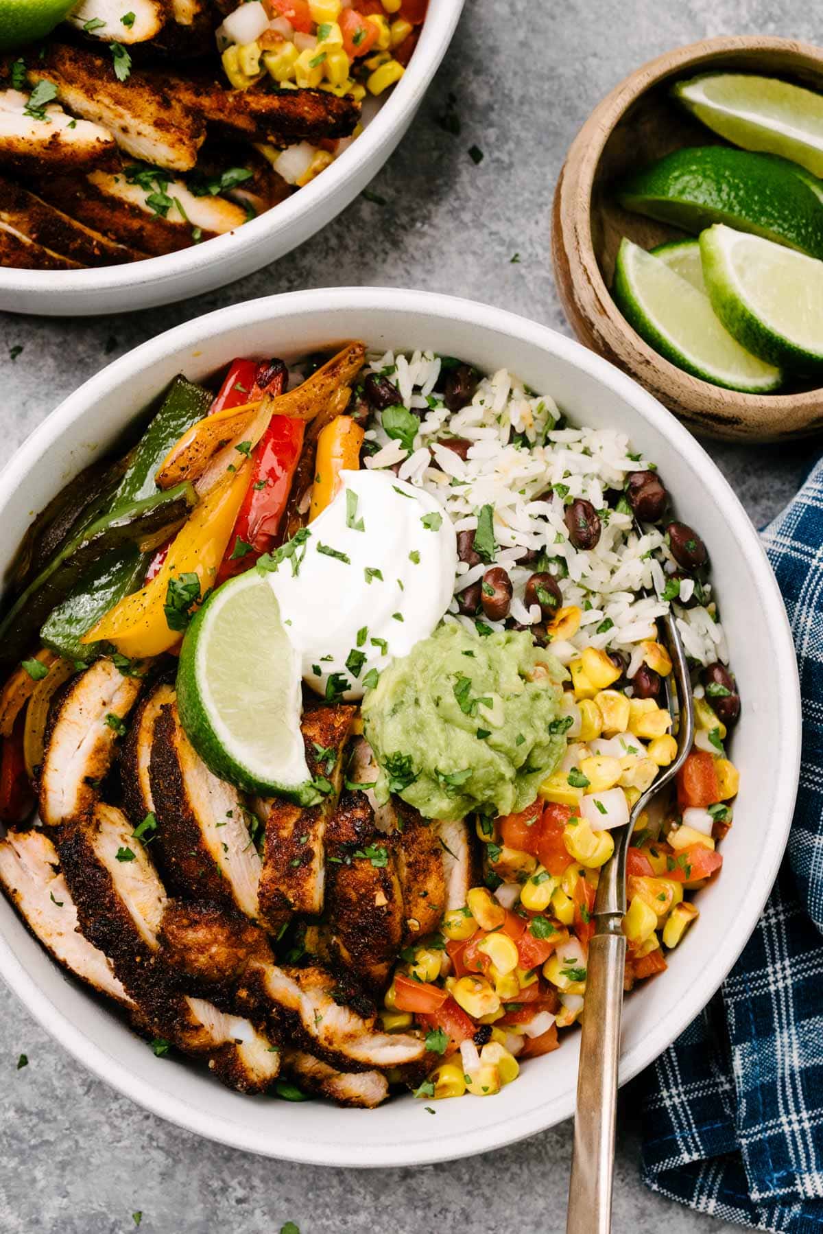 A fork tucked into a chicken fajita bowl with seared chicken, sauteed onions and peppers, black beans and rice, and corn salsa; a second fajita bowl, small bowl of lime wedges, and blue napkin surround the bowl.