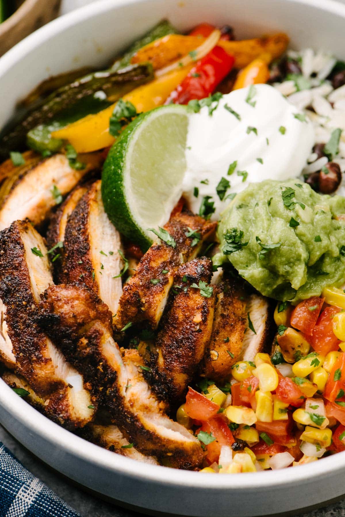 Side view, a close up view of a chicken fajita bowl with sliced fajita chicken, sautéed peppers and onions, tomato corn salsa, and black beans and rice; garnished with sour cream, guacamole, and a lime wedge.