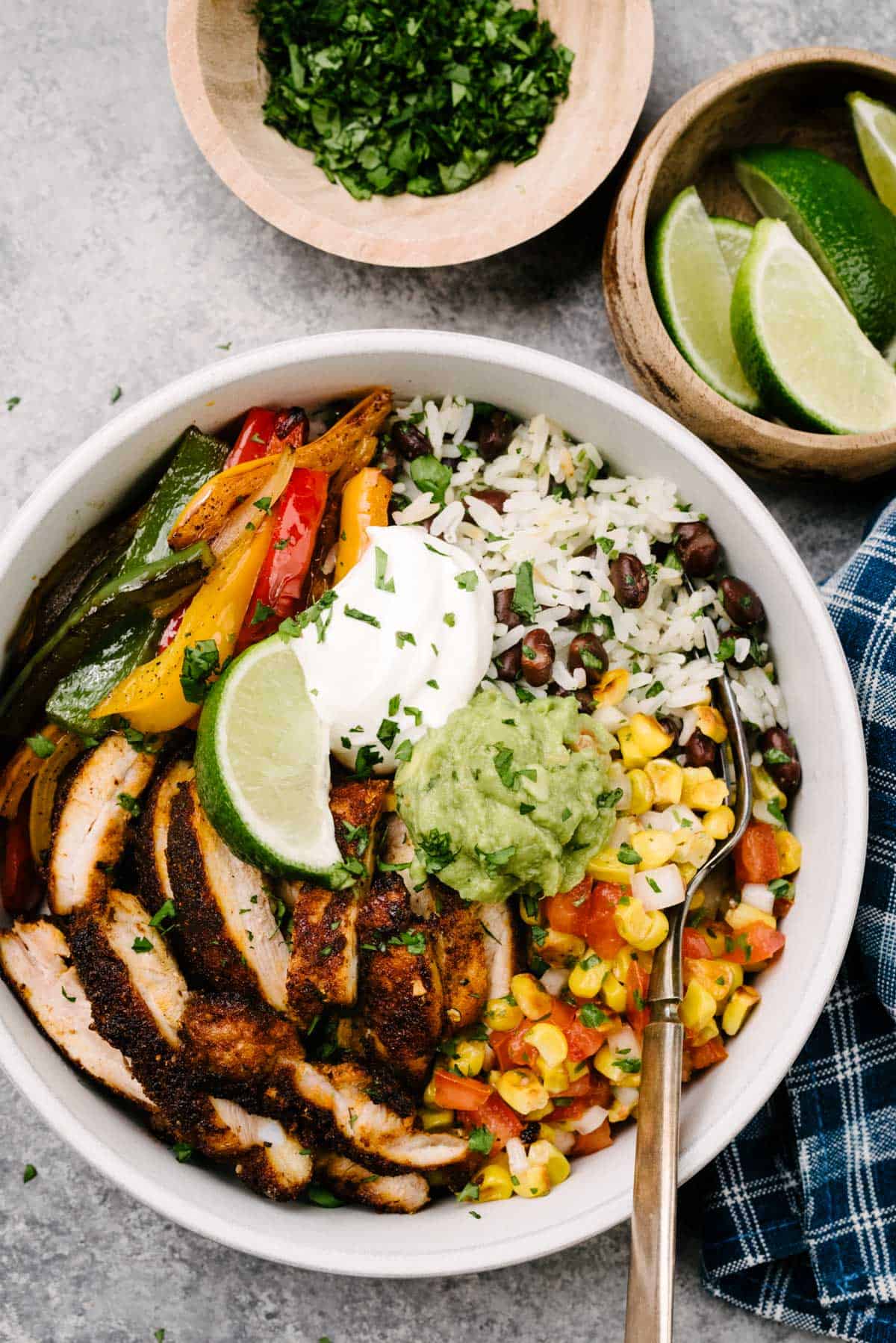 A fork tucked into a chicken fajita bowl with seared chicken, sauteed onions and peppers, tomato corn salsa, and black beans and rice; topped with guacamole, sour cream, and a lime wedge. Small bowls of cilantro and lime wedges as well as a blue napkin surround the bowl.