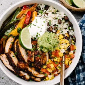 A silver fork tucked into a chicken fajita bowl; the fajita bowl is made with thinly sliced fajita chicken, sauteed onions and peppers, tomato corn salsa, and black beans and rice, garnished with a dollop of sour cream, guacamole, and a lime wedge.