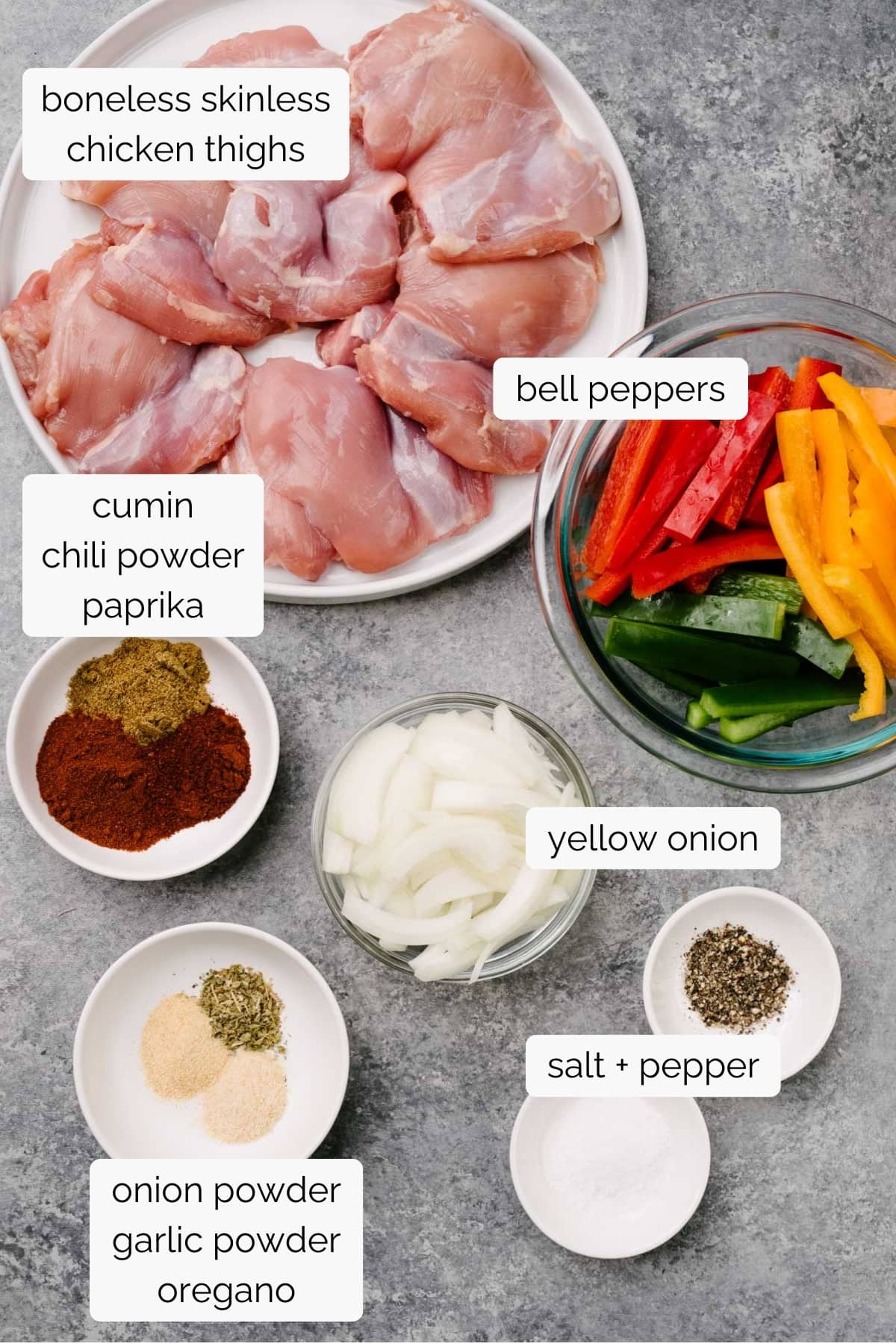 The ingredients for chicken and veggie fajitas arranged in small bowls on a concrete background - boneless skinless chicken thighs, bell peppers, yellow onion, olive oil, and fajita seasonings.