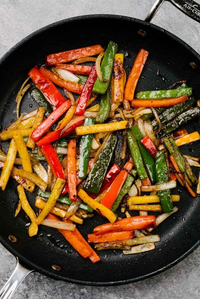 Thinly sliced sautéed onions, green bell pepper, red bell pepper, and yellow bell pepper in a black skillet.