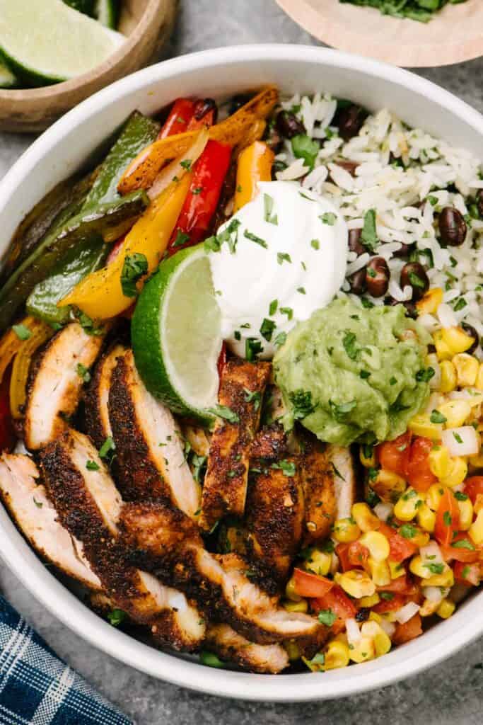 A chicken fajita bowl with fajita chicken, sauteed peppers and onions, black beans and rice, and tomato corn salsa in a low white bowl, garnished with sour cream, guacamole, and lime wedge.