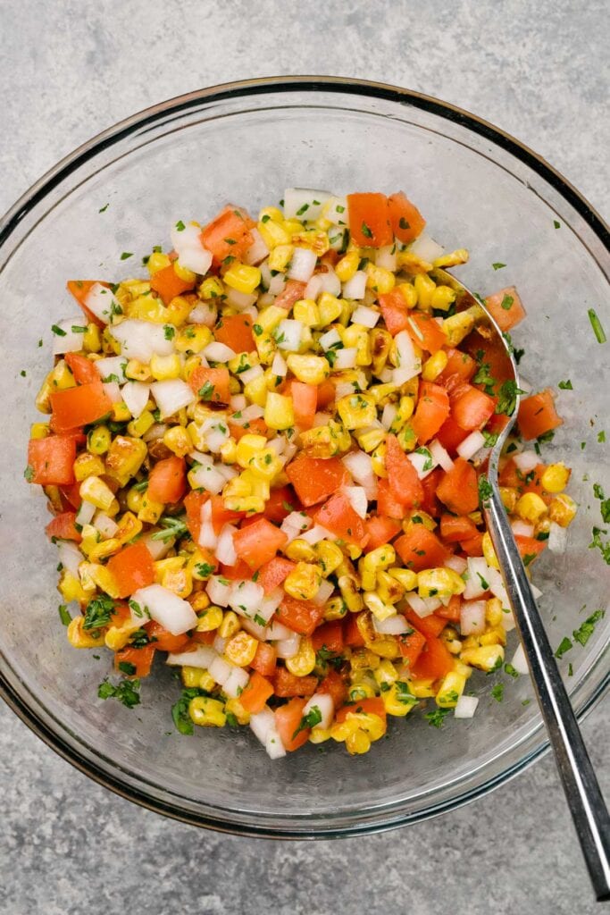 A spoon tucked into a glass mixing bowl filled with tomato corn salsa. 