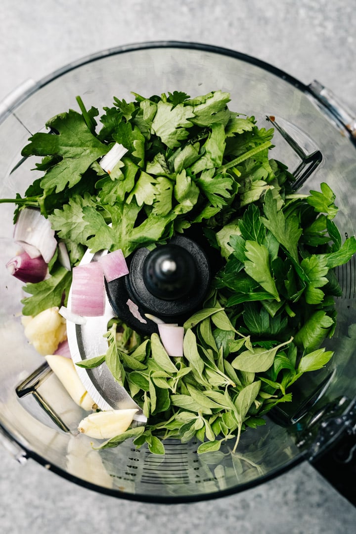 Cilantro, parsley, oregano, garlic, and shallot in the bowl of a food processor.