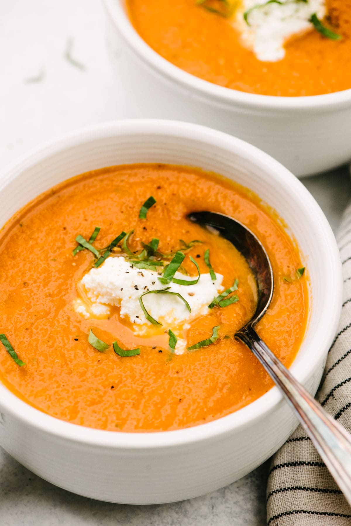 Side view, two bowls of creamy tomato basil soup garnished with ricotta cheese and fresh basil.