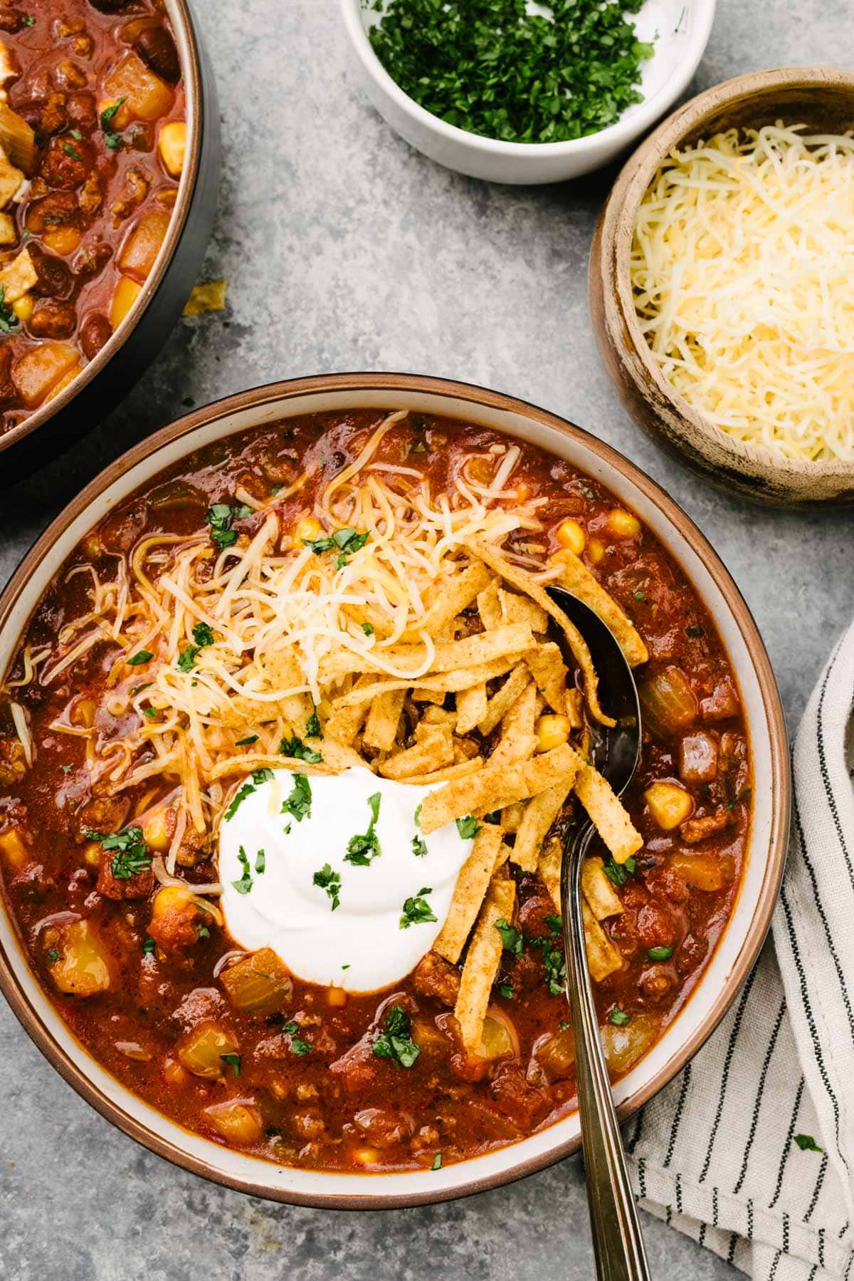 A spoon tucked into a bowl of crockpot taco soup, garnished with sour cream, shredded cheese, tortilla strips, and chopped cilantro; the bowl is surrounded by a striped linen napkin and small bowls of shredded cheese and cilantro.