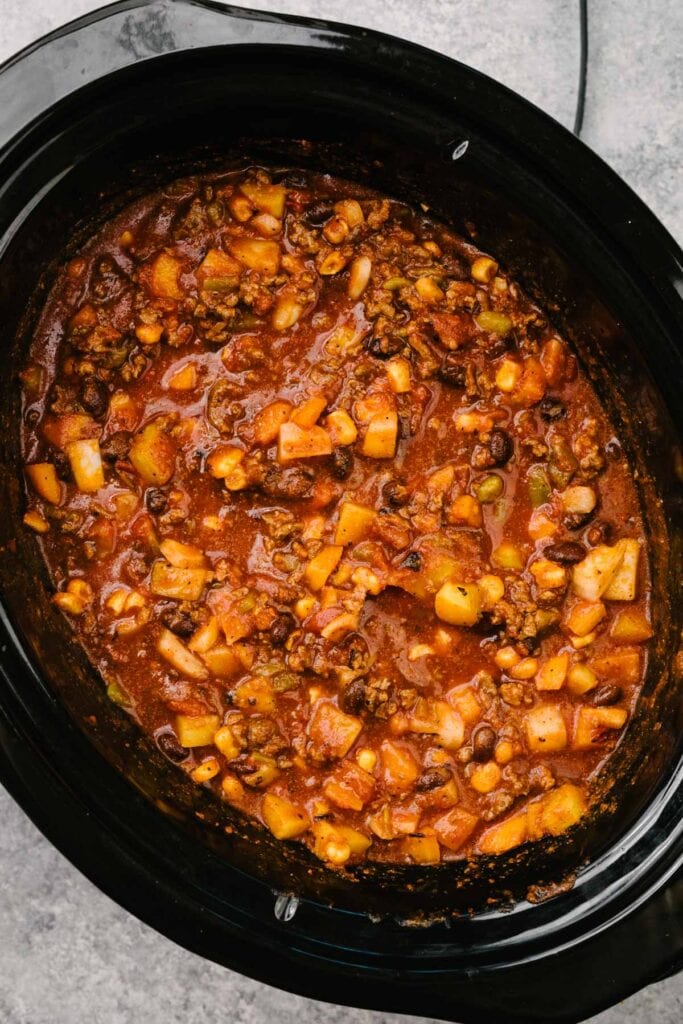 Cooked ground beef mixed with taco seasoning, tomato paste, onion, bell pepper, crushed tomatoes, diced green chiles, black beans, frozen corn, and beef broth in a crockpot before slow cooking.