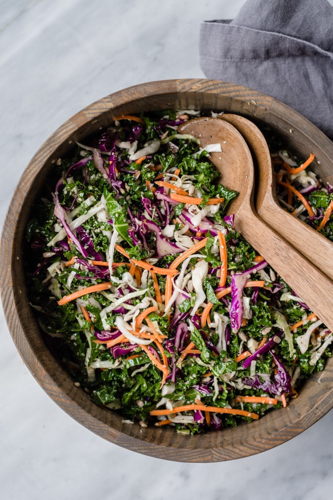 Crunchy kale slaw (or salad) tossed with red and green cabbage, carrots, hemp seeds, and sunflower seeds in a wood serving bowl.