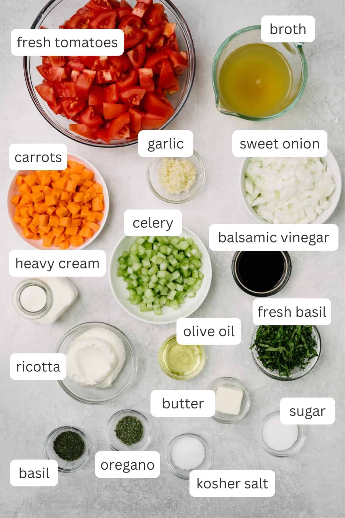 The ingredients needed to make fresh tomato soup arranged in small bowls on a concrete background. The ingredient list includes fresh tomatoes, broth, onions, carrot, celery, garlic, balsamic vinegar, heavy cream, fresh basil, dried basil, dried oregano, sugar, salt, pepper, olive oil, butter, and ricotta cheese.