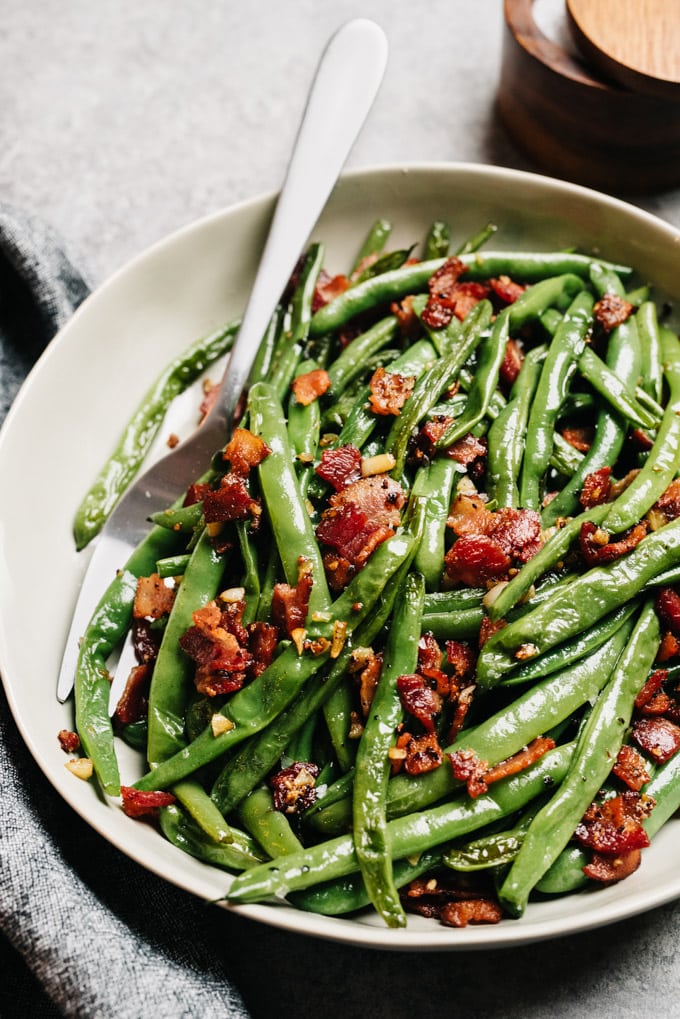 Side view, green beans with bacon in a tan serving bowl with a silver serving fork.