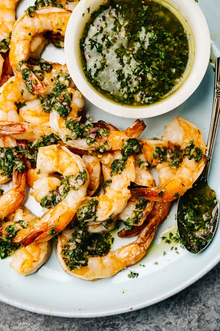 Grilled shrimp drizzled with chimichurri sauce on a blue serving plate with a small bowl of sauce to the side.