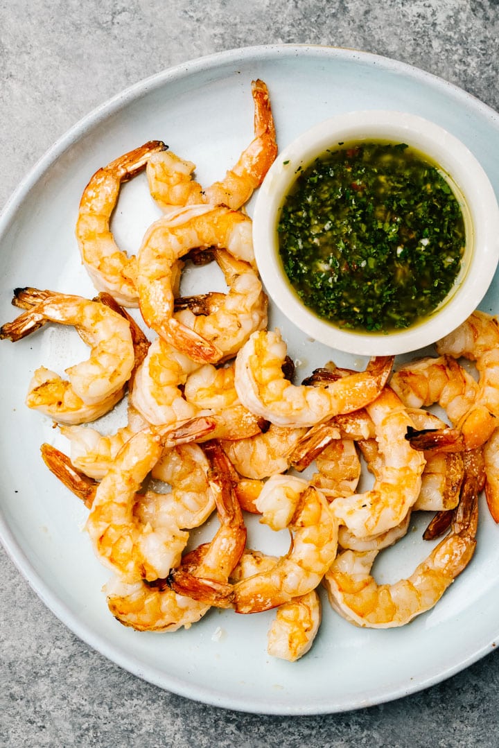 Grilled shrimp on a blue serving platter with a small dip bowl filled with chimichurri sauce.