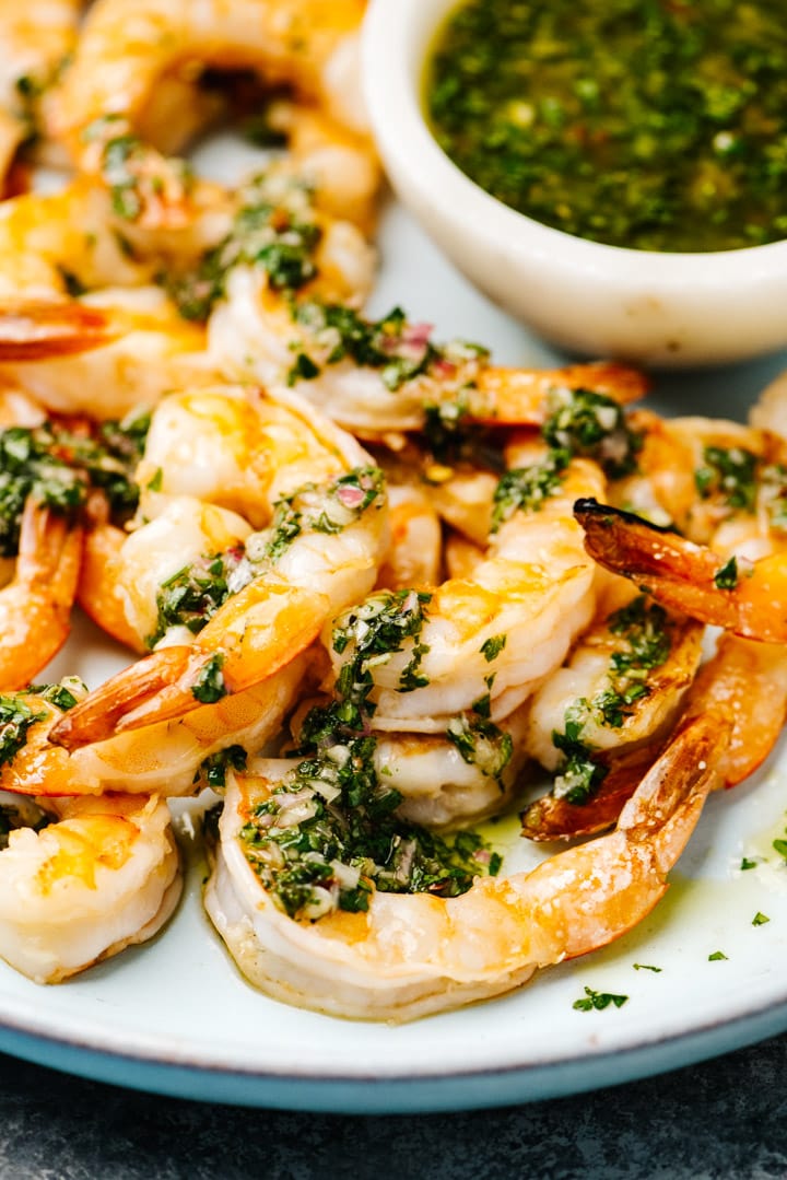 Side view, grilled shrimp drizzled with chimichurri sauce on a blue serving platter.