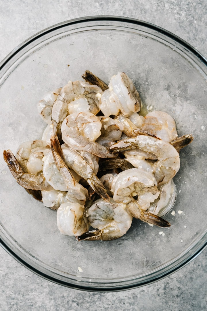 Raw shrimp in a glass bowl tossed with olive oil, lemon juice, garlic, and salt.