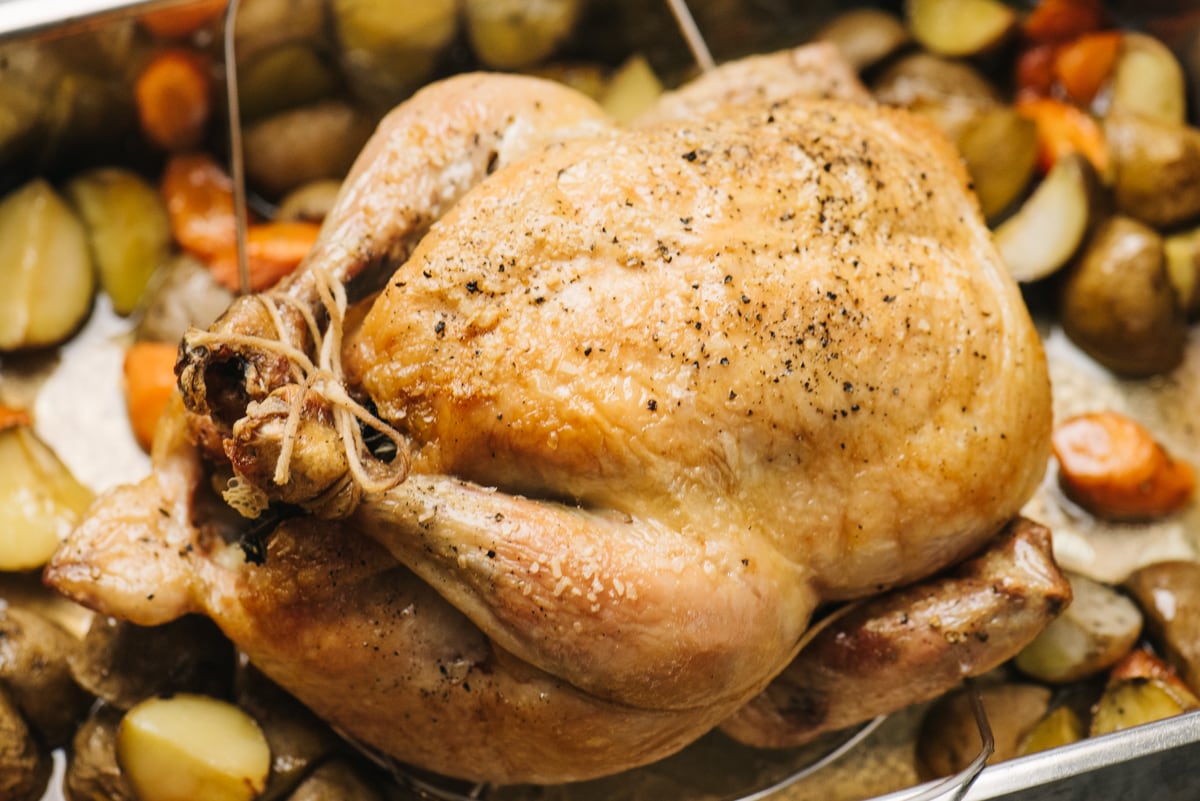 A whole roasted chicken in a roasting pan surrounded by oven roasted potatoes and carrots.