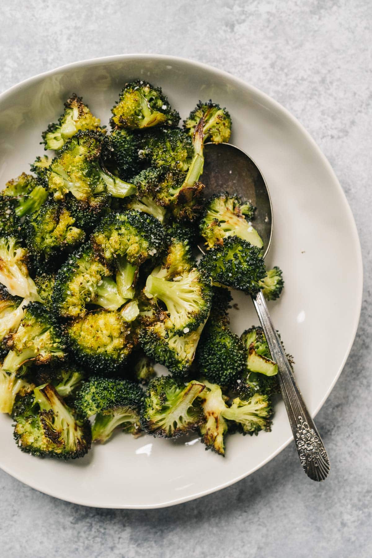 Oven roasted broccoli in a tan serving bowl with a silver serving spoon on a concrete background.