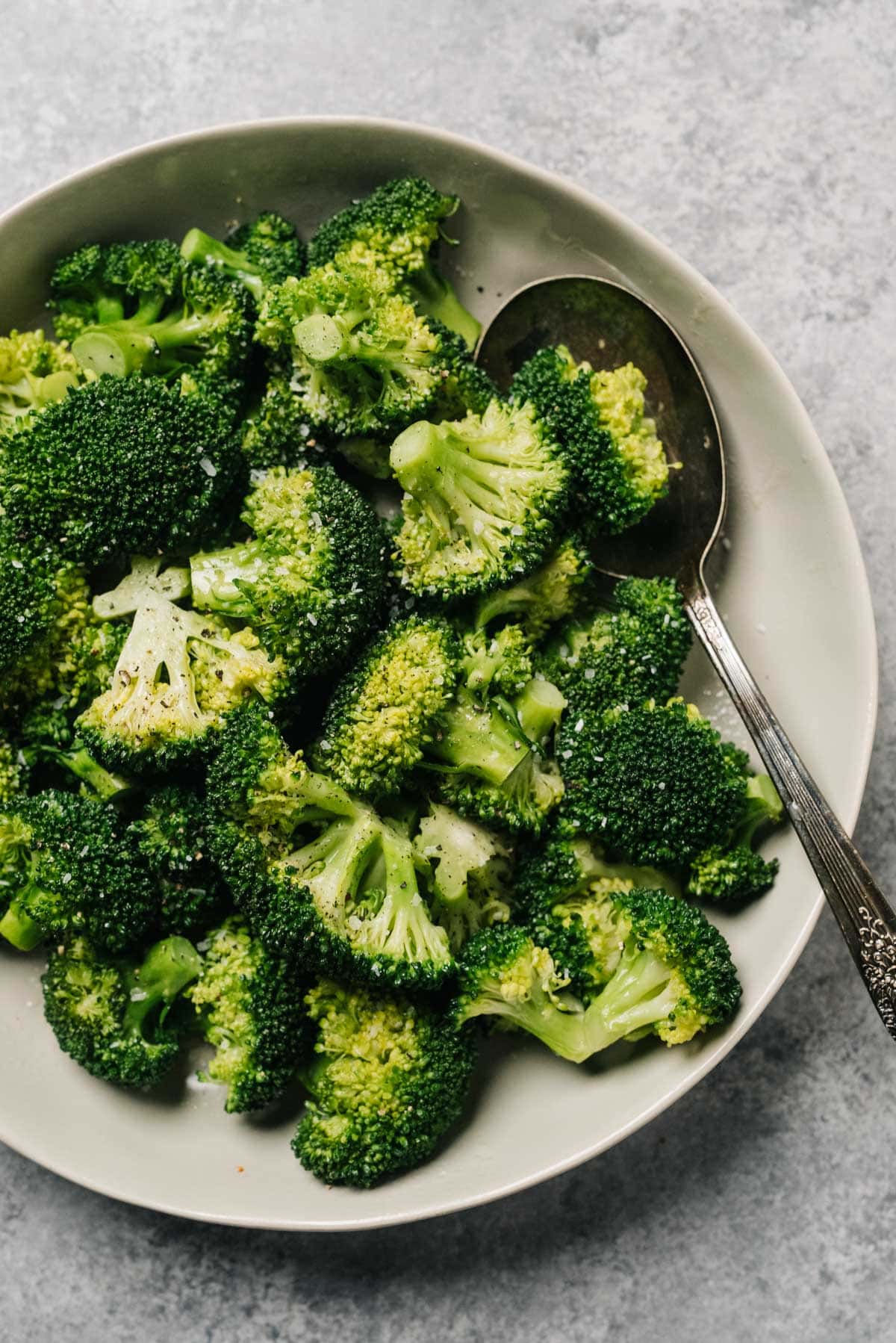 Steamed broccoli in a tan serving bowl with a silver serving spoon on a concrete background.