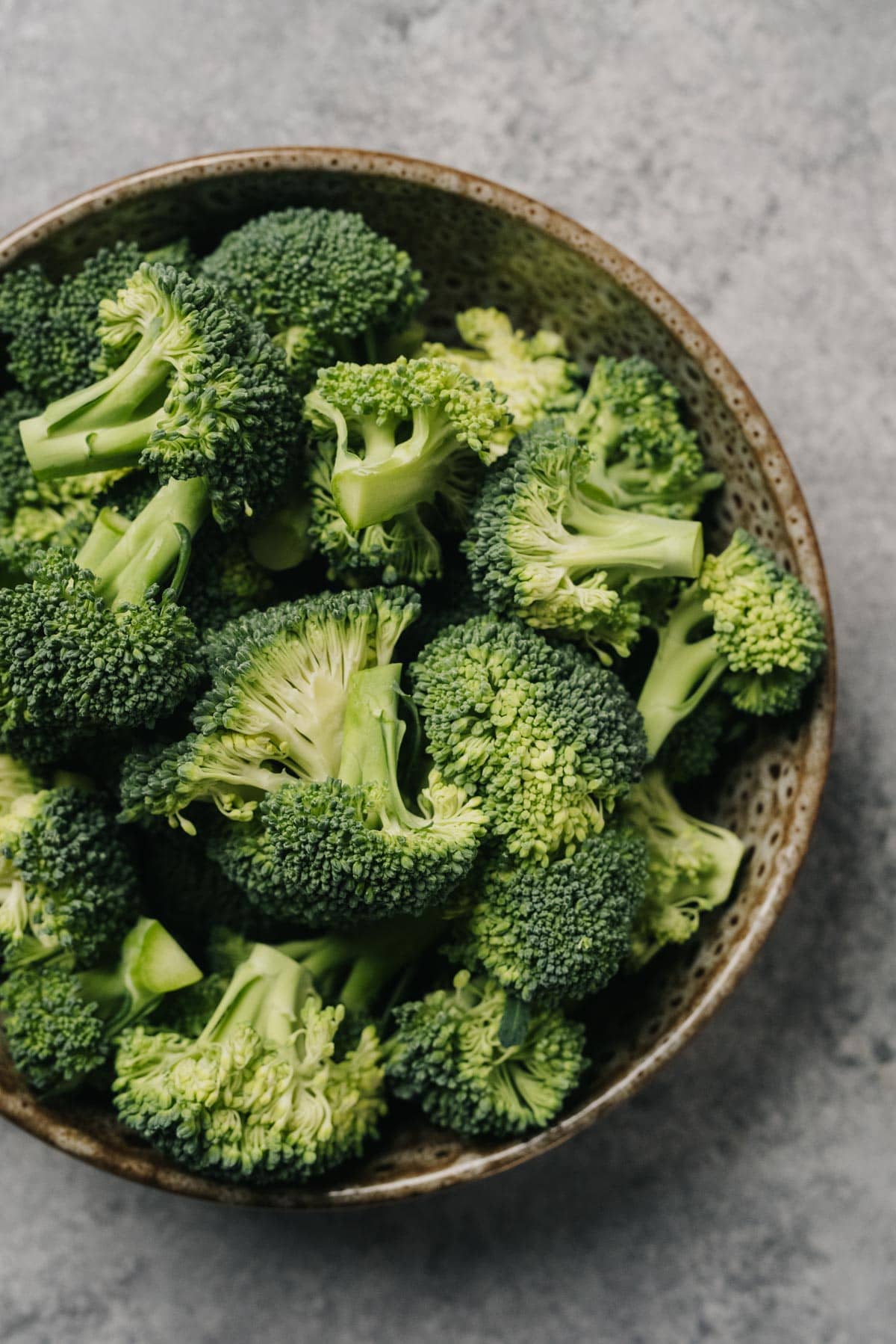 Raw broccoli florets in a brown speckled bowl on a concrete background.