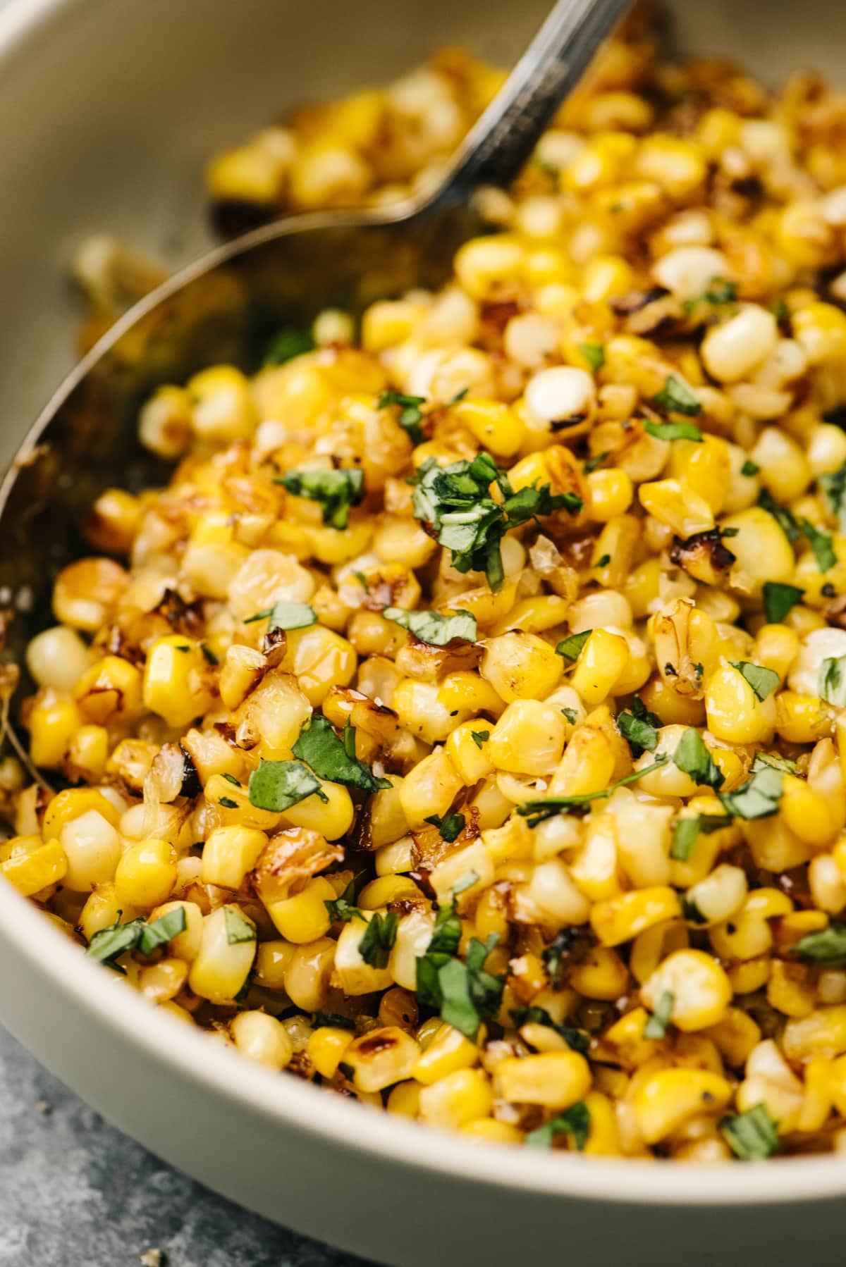 Side view, sauteed fresh corn in a tan bowl with fresh herbs and silver serving spoohn. 