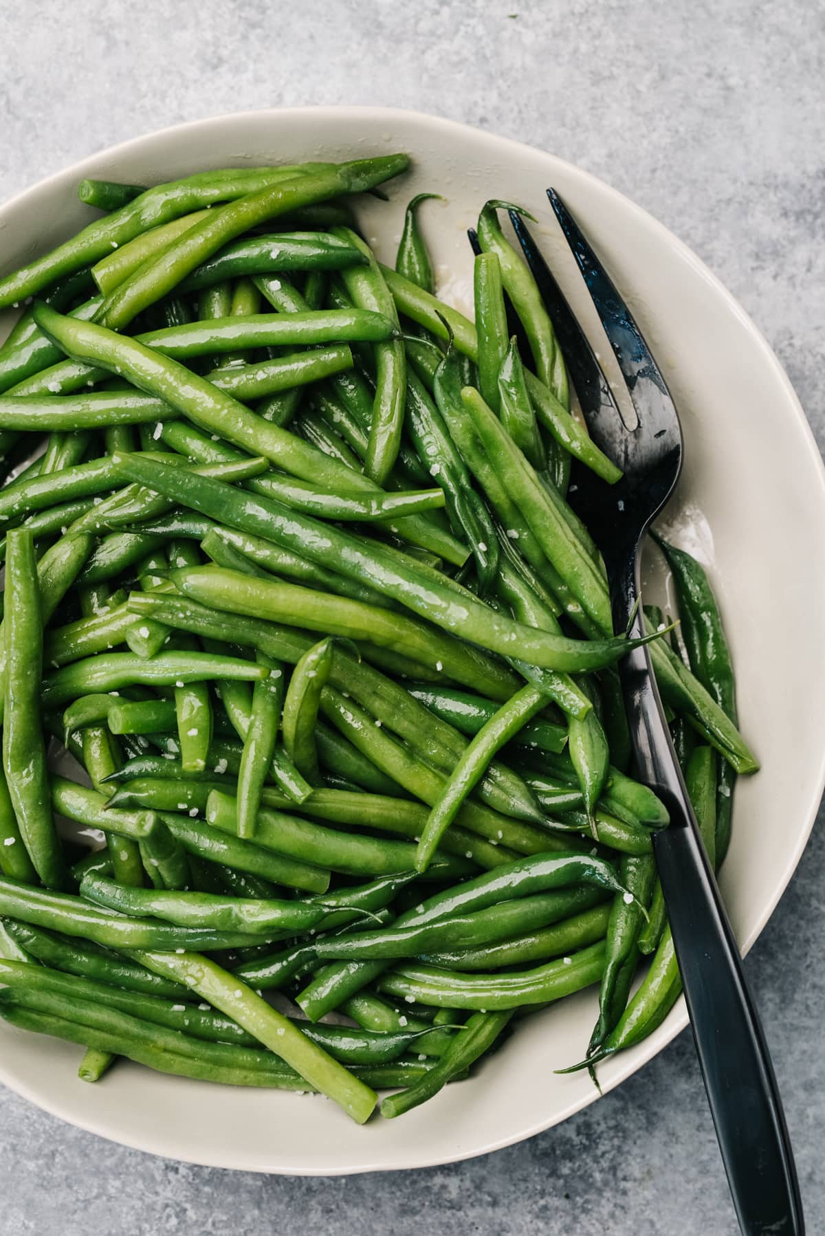 Steamed green beans in a tan serving bowl with a black serving fork.