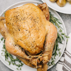 A whole roasted chicken on a white platter surrounded with fresh herbs for garnish.