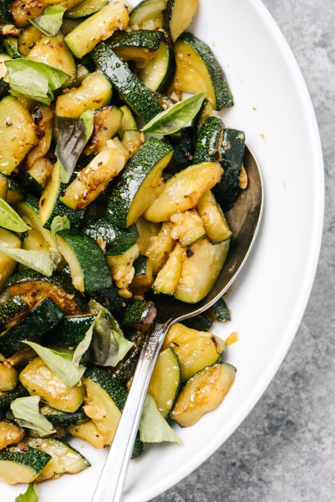 Sautéed zucchini in a white bowl with a silver serving spoon.