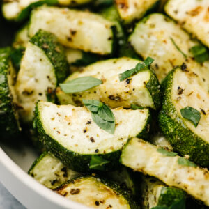 Side view, roasted zucchini in a tan serving bowl, garnished with fresh basil.