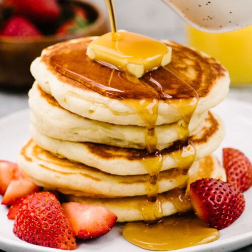 Side view, pouring maple syrup from a small speckled pitcher over a stack of fluffy homemade pancakes topped with butter and plated with fresh strawberries.