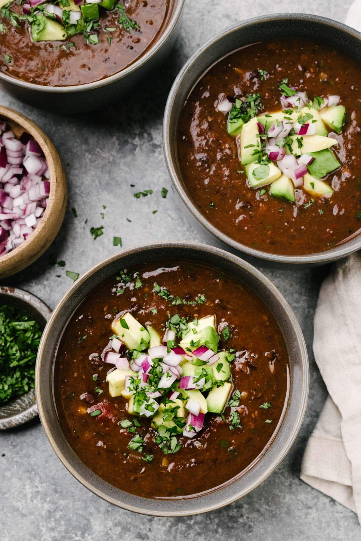 Three bowls of Instant Pot Black Bean Soup on a concrete surface, garnished with diced avocado, minced red onion, and fresh cilantro; the bowls are surrounded by a cream colored linen napkin and small bowls of red onion and cilantro.