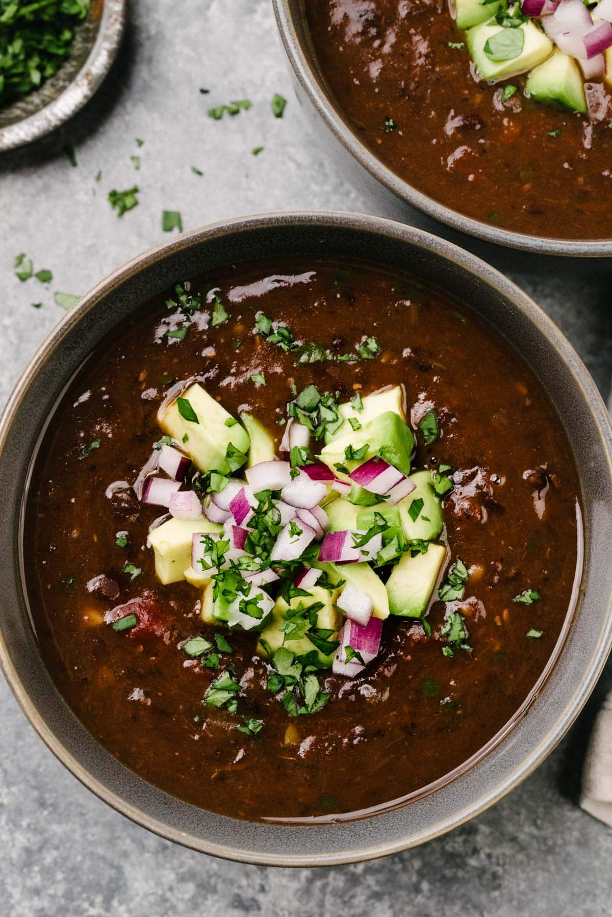 A bowl of Instant Pot black bean soup, garnished with diced avocado, minced red onion, and fresh cilantro; another bowl of soup and a small bowl of chopped cilantro surround the bowl.