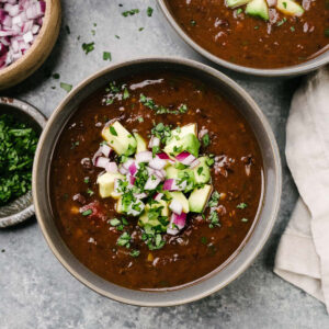 A bowl of Instant Pot black bean soup on concrete background garnished with diced red onion, diced avocado, and chopped cilantro; a tan linen napkin and two bowls of cilantro and avocado surround the bowl.