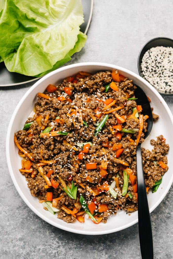 Ground beef lettuce wrap filling in a white bowl with black spoon with a platter of lettuce leaves and small bowl of sesame seeds to the side.