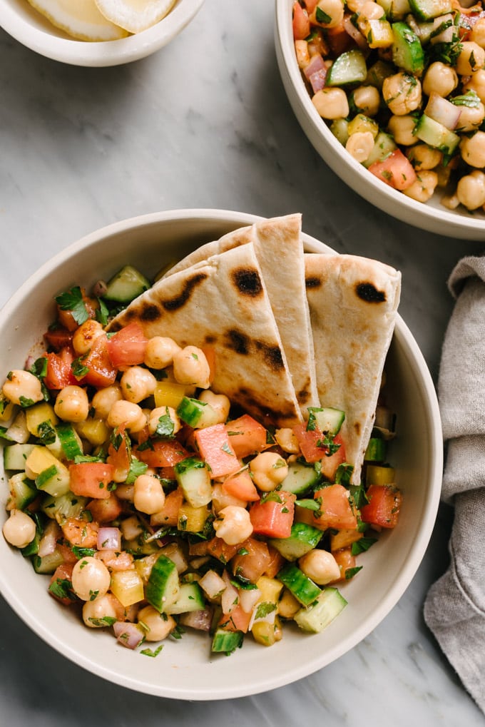 Two bowls of mediterranean chickpea salad on a marble table with a linen napkin and small dish of lemon wedges.