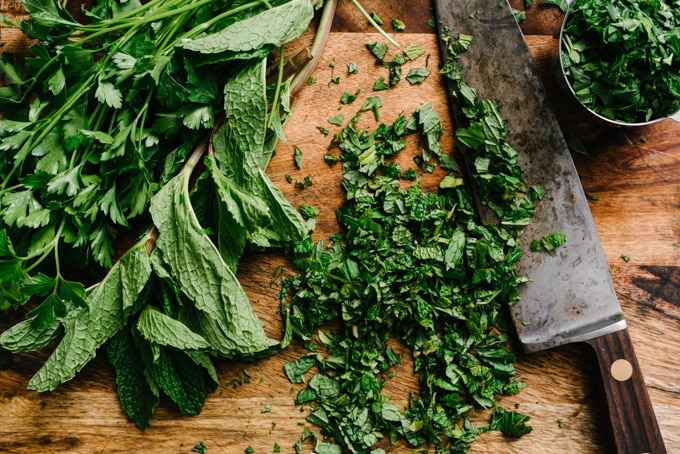 Minced mint on a cutting board next to bundles of parsley and mint.