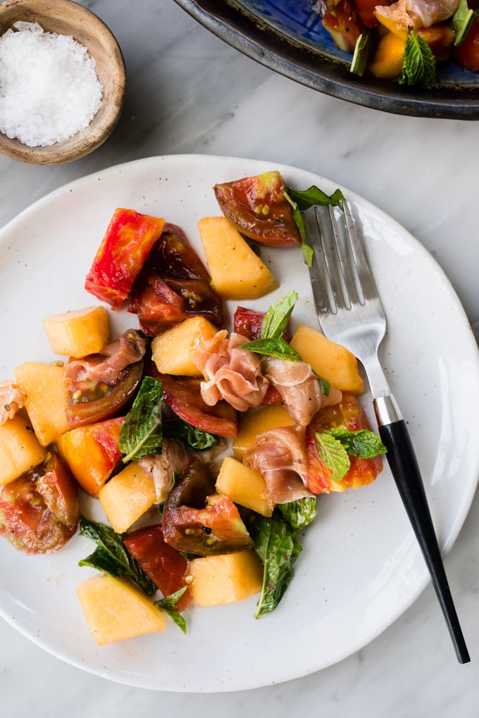 Melon salad with prosciutto, tomato, mint, and basil on a white plate with a fork.