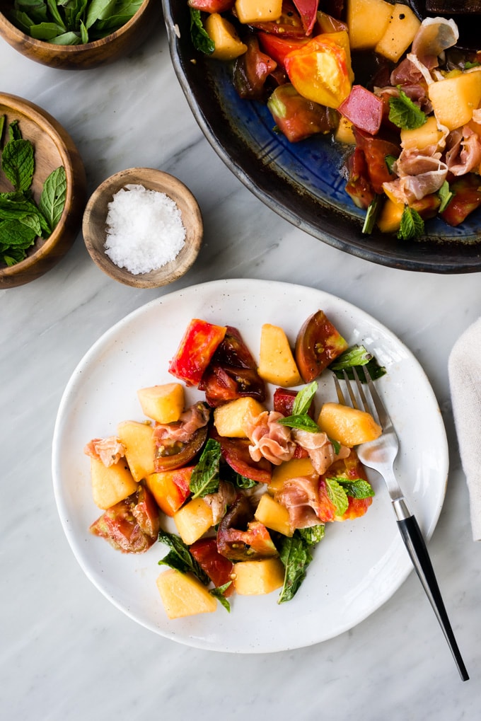 A plate of summer melon salad with prosciutto on a marble table with small dishes of mint, basil, and sea salt.