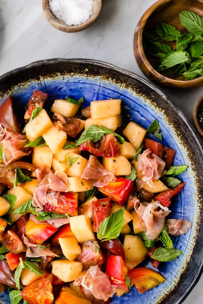 Melon salad with prosciutto, tomato, mint, and basil on a marble table.