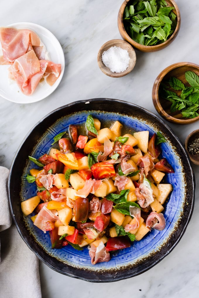 Summer melon salad with prosciutto, tomato, mint, and basil tossed with apple cider vinaigrette in a blue salad bowl on a marble table.