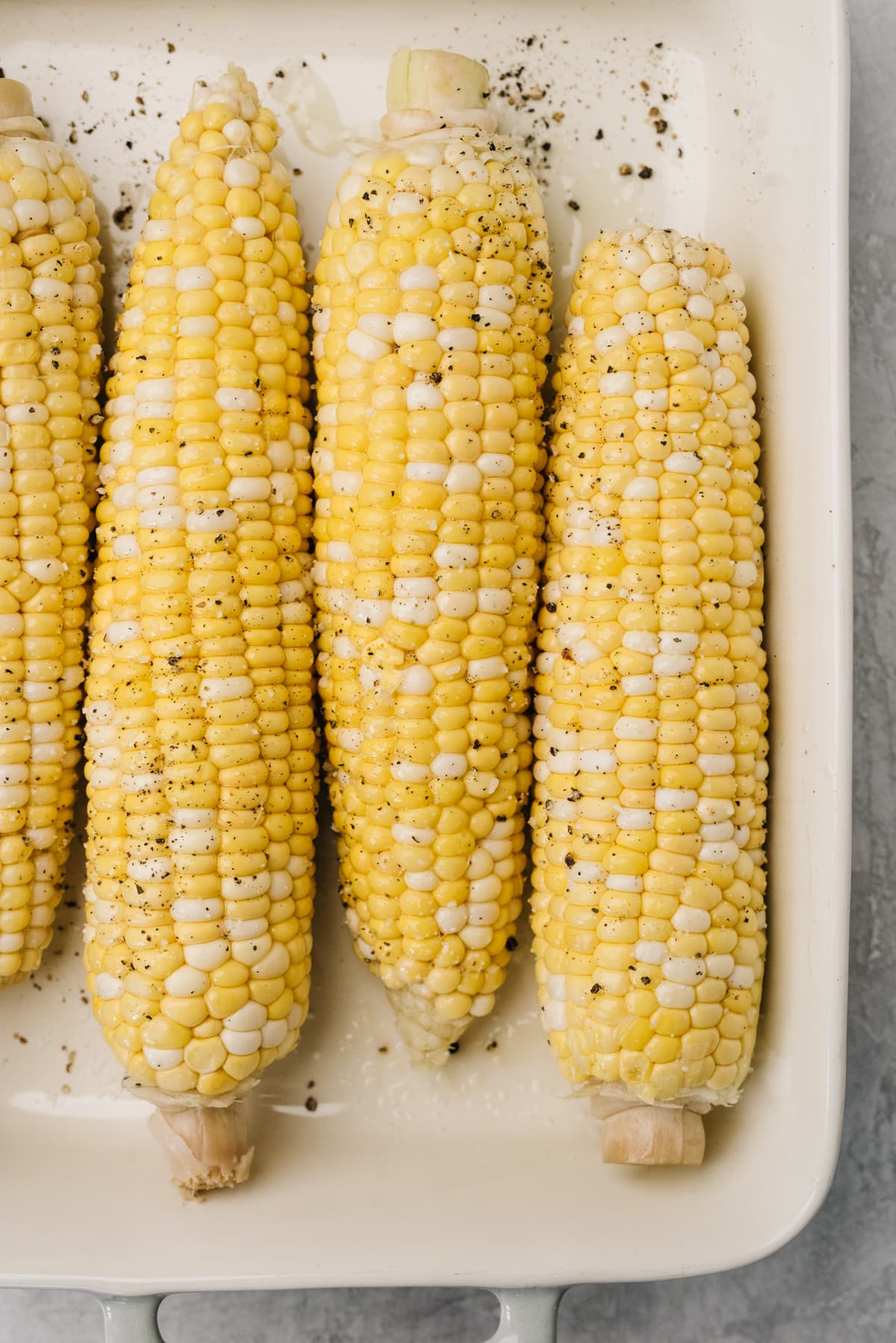 Four ears of corn with husks and silks removed, drizzled with olive oil, salt, and pepper, in a casserole dish.