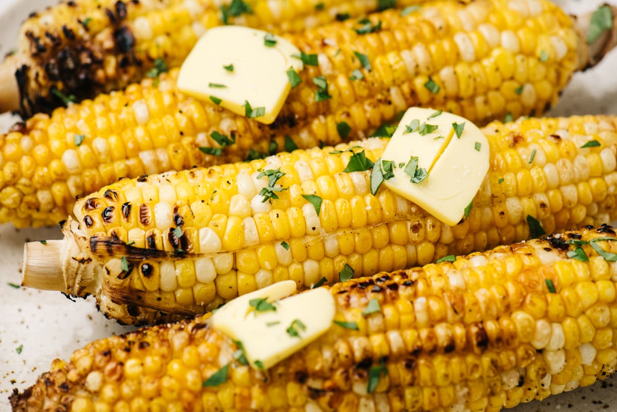 Side view, four ears of grilled corn on the cob topped with butter and chopped basil.